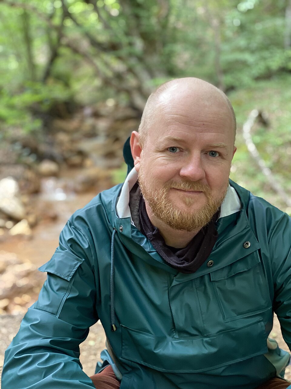 Upper body picture of Ray Nayler smiling towards the camera, wearing a green waterproof jacket, with woodlands in the background.