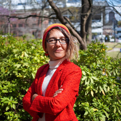 Upper body shot of Natalie Khazaal smiling towards the camera with bushes, a tree and buildings in the background.